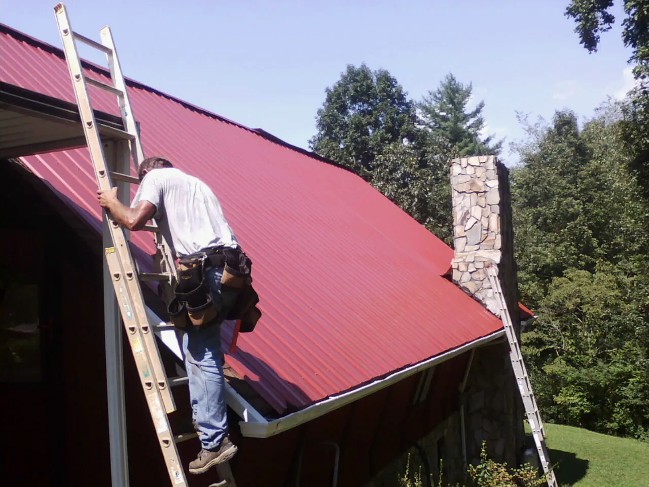 Roofer on a ladder beside a red metal roof and stone chimney — Soffit & Fascia Repair by Roofing Brothers in Jourdanton